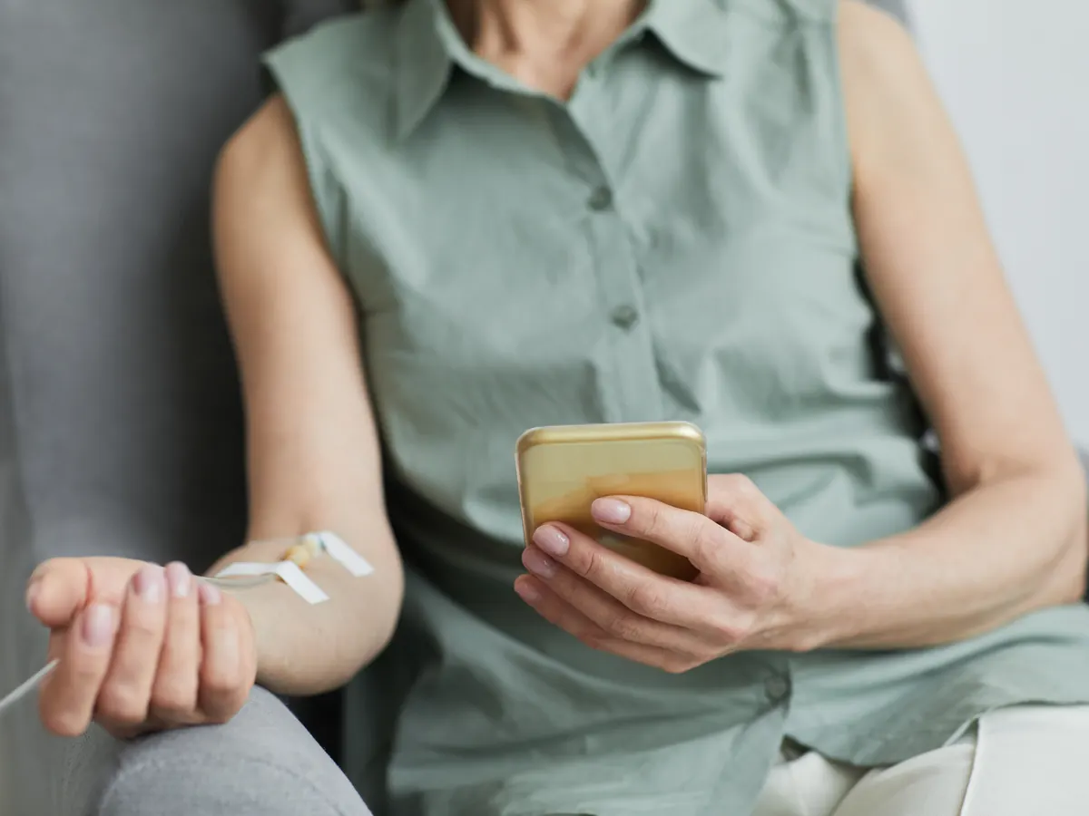 A woman checks her phone while receiving IV fluids in an armchair at home