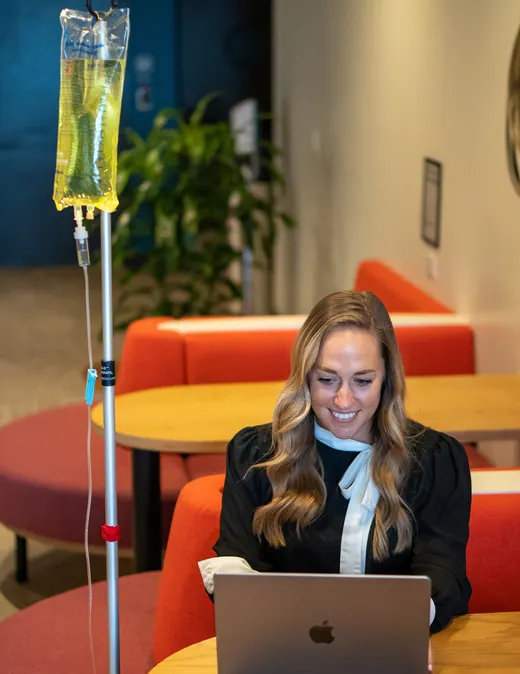 A client works on her laptop while an IV drip runs during a home visit